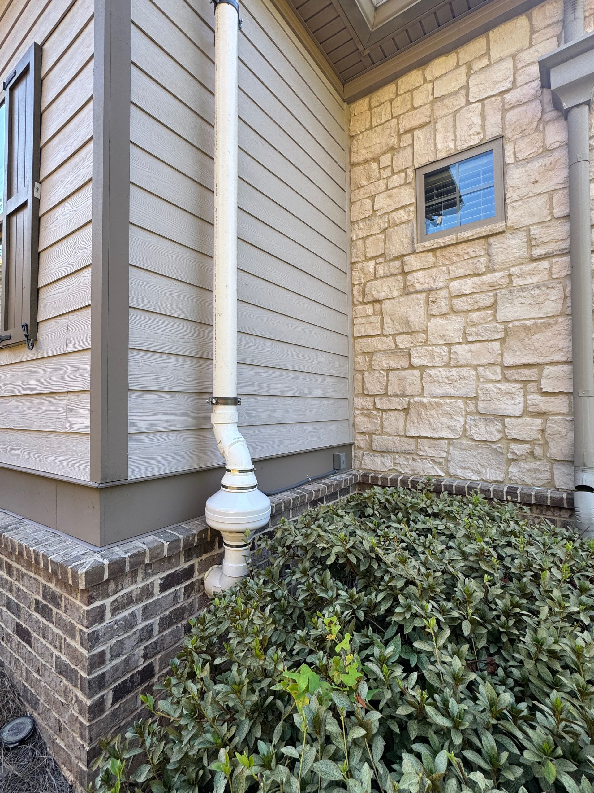 White downspout on a house with gray siding and brick facade, emptying into nearby shrubs.