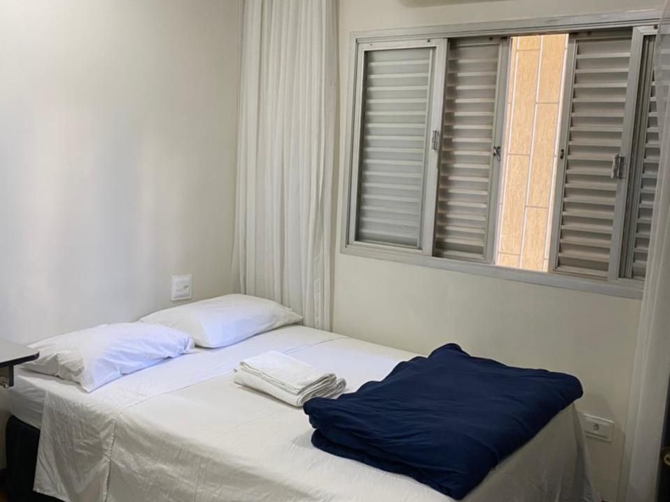 Um quarto arrumado com uma cama com lençóis brancos, dois travesseiros, toalhas brancas dobradas e uma manta azul-escura dobrada.