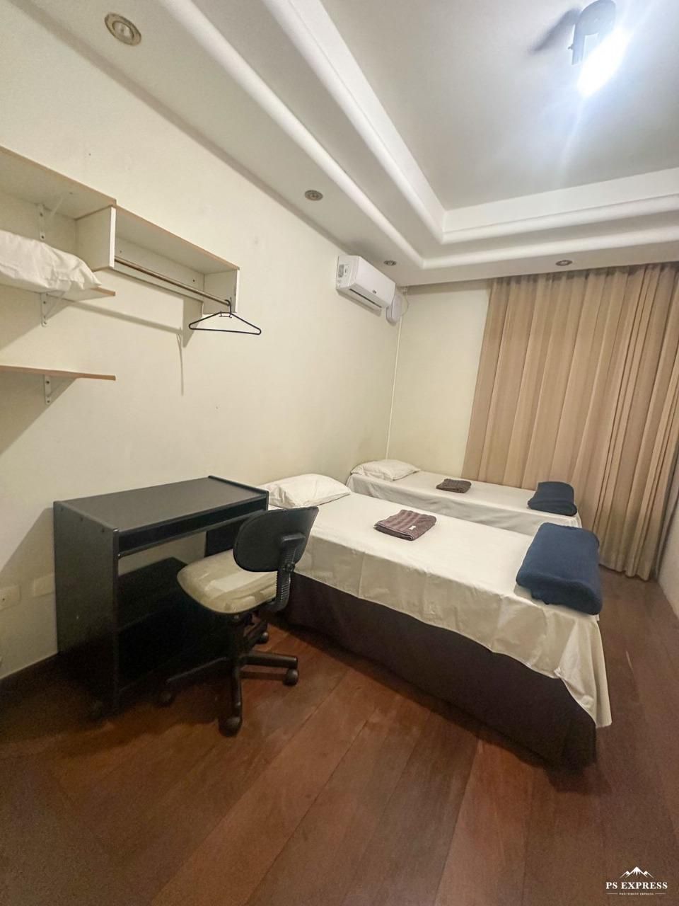 Um quarto de hotel com duas camas de solteiro, uma escrivaninha preta, uma cadeira e prateleiras fixadas na parede sobre um piso que imita madeira.