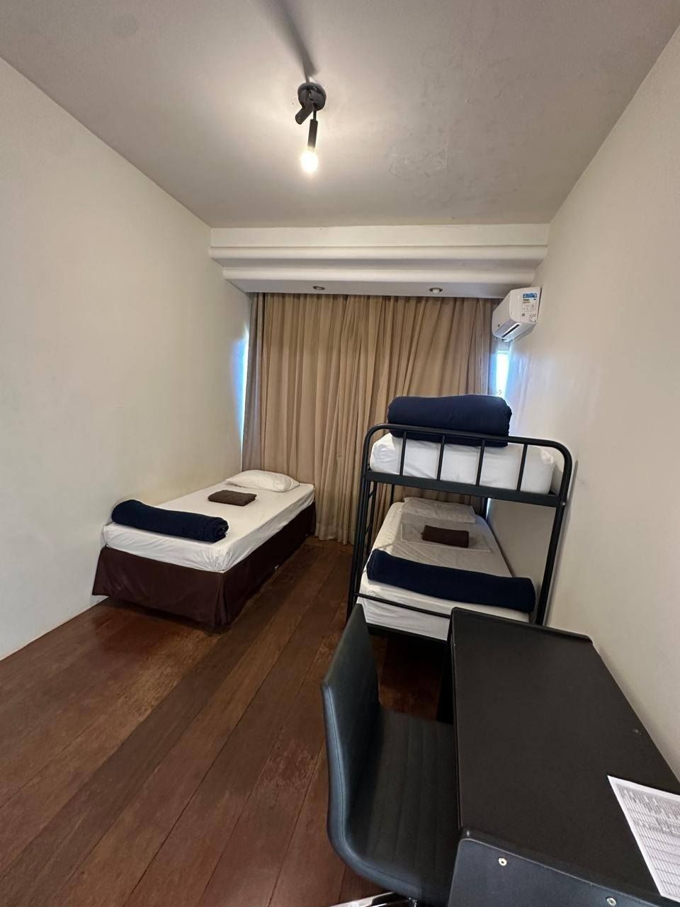 Um pequeno quarto de hotel em tons neutros, com uma cama de solteiro, um beliche de metal preto, piso de madeira e uma escrivaninha com cadeira.