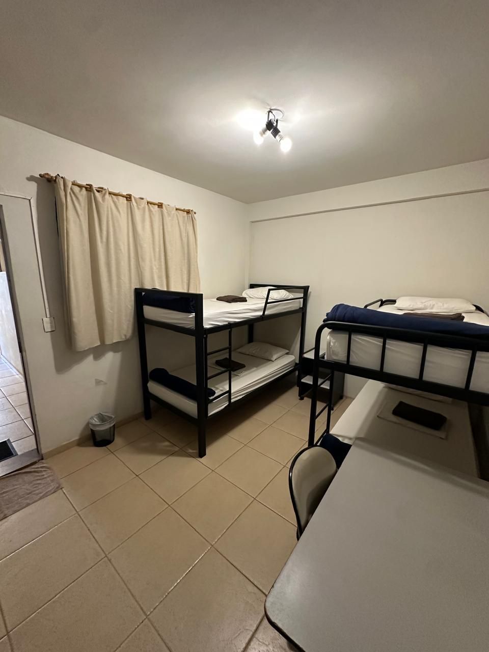 Um quarto simples com um beliche, uma cama de solteiro elevada, uma escrivaninha, uma cadeira e piso de azulejo claro.
