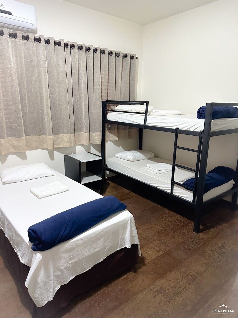 Um quarto de hotel com uma cama de solteiro e um beliche, com lençóis brancos, detalhes em azul-marinho e cortinas bege.
