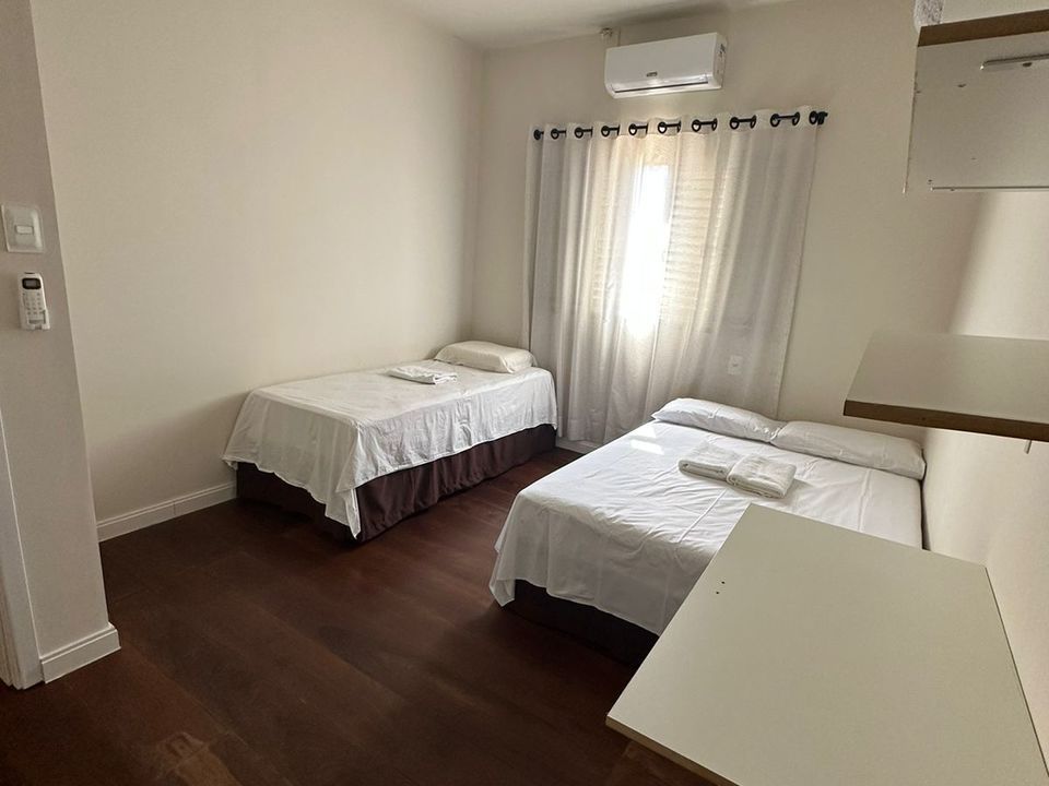 Um quarto com piso de madeira escura, uma cama de solteiro, uma cama maior com lençóis brancos e um ar-condicionado acima da janela.