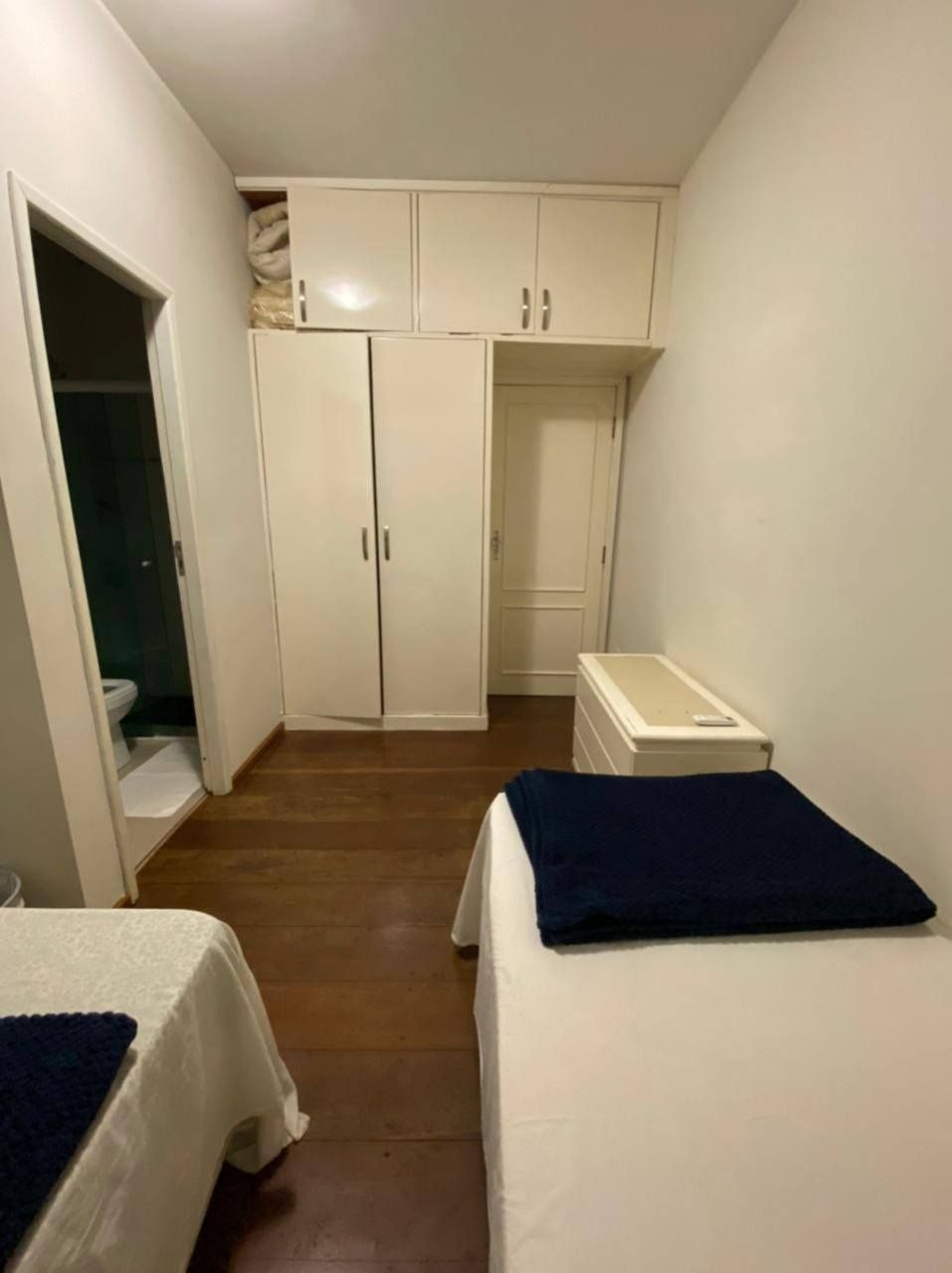Um pequeno quarto com duas camas de solteiro, piso de madeira, armários embutidos brancos e uma porta que dá acesso a um banheiro.