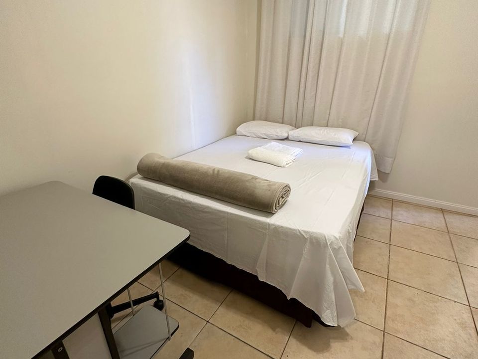 Uma cama com lençóis brancos e um cobertor cinza, ao lado de uma escrivaninha e cadeira cinzas, em um quarto com piso de azulejo claro.