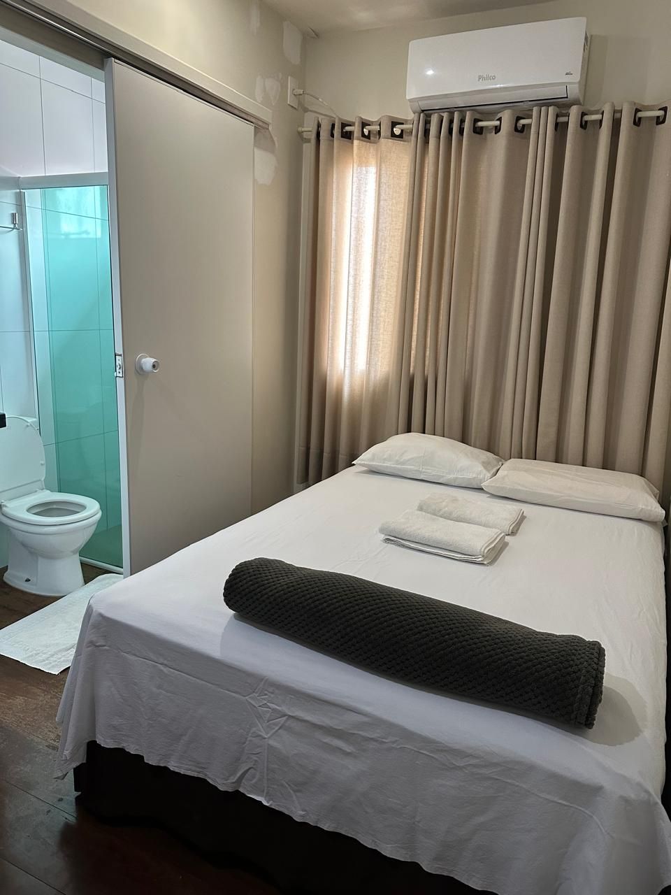 Um quarto de hotel com uma cama com lençóis brancos e um travesseiro cilíndrico escuro, ao lado de um banheiro com vaso sanitário.