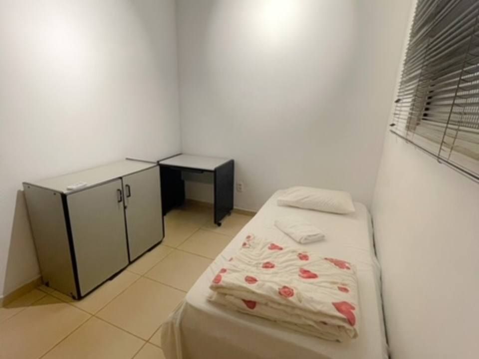 Um quarto compacto com uma cama de solteiro com edredom estampado, um pequeno armário cinza e uma escrivaninha encostada em uma parede branca.