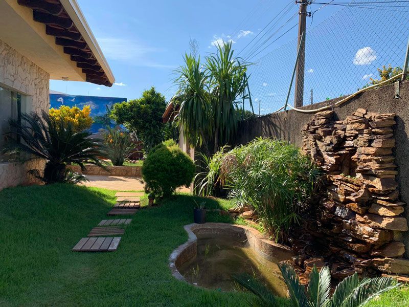 Um jardim nos fundos da casa com uma cascata de pedra, um pequeno lago, um gramado exuberante com caminho de lajes de madeira e plantas tropicais.