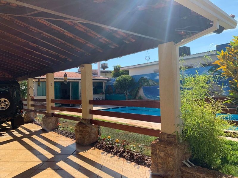 Uma pérgola de madeira oferece vista para um quintal ensolarado com piscina e uma casa com uma parede pintada de azul.