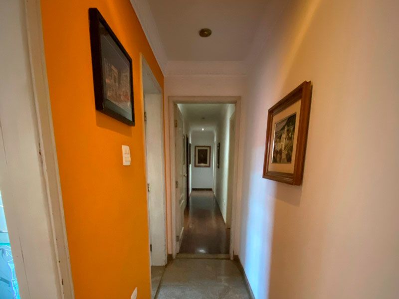 Um corredor estreito com uma parede esquerda laranja vibrante, uma parede direita creme, dois quadros emoldurados e uma porta no final.