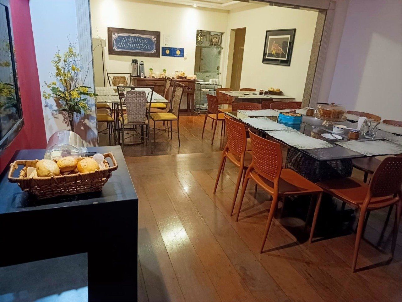 Uma sala de jantar bem iluminada apresenta uma grande mesa com cadeiras laranja, piso de madeira e uma cesta de pão na entrada.