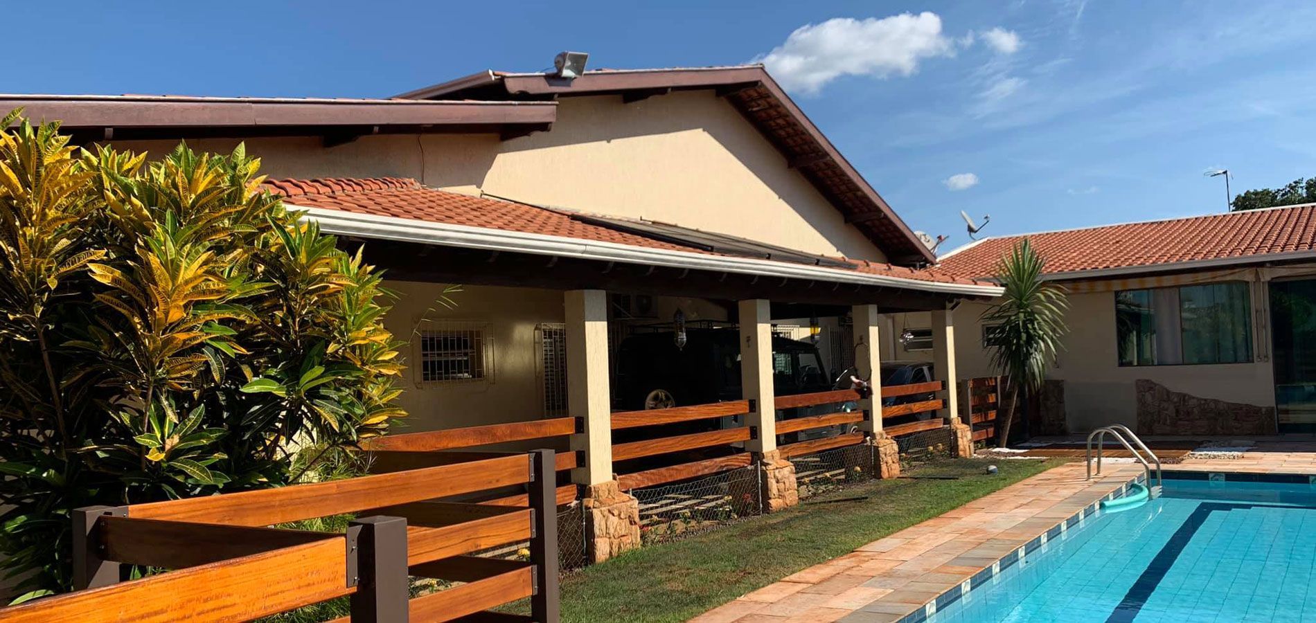 Uma casa estilo rancho com varanda de madeira e telhado de telhas ao lado de uma piscina azul sob um céu ensolarado.