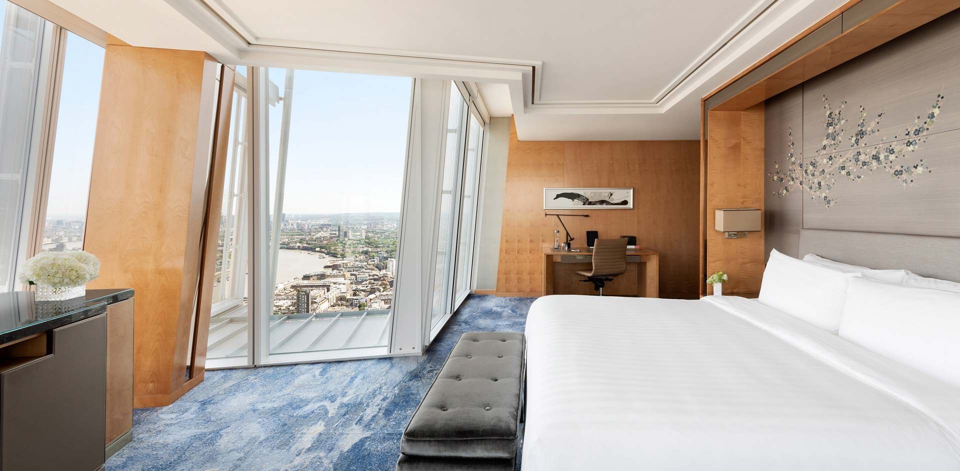 Um quarto de hotel moderno e sofisticado com uma grande cama branca, janelas do chão ao teto com vista para a paisagem urbana e painéis de madeira.