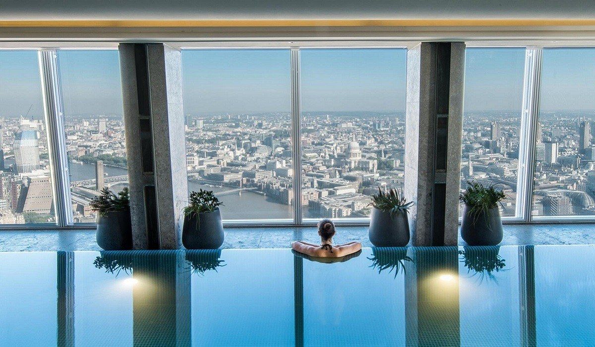 Uma pessoa relaxa em uma piscina infinita coberta, com vista para o horizonte da cidade através de grandes janelas que vão do chão ao teto.