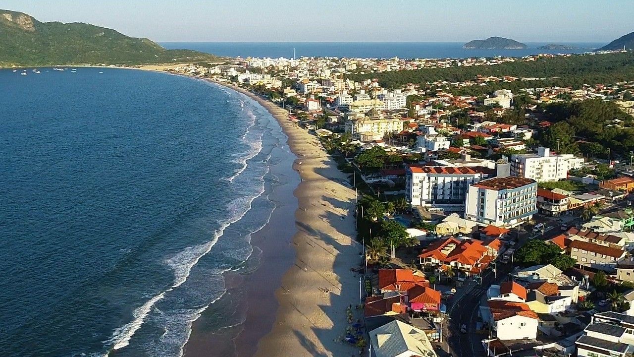 Vista aérea de uma praia de areia que acompanha uma cidade costeira com edifícios, palmeiras e águas azuis do oceano.