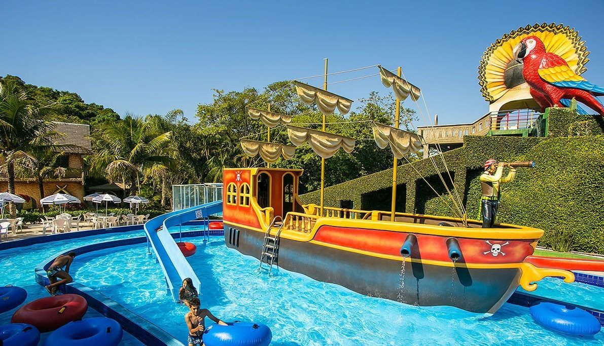 Uma peça central em forma de navio pirata fica em um rio artificial de um parque aquático, com boias azuis, um toboágua e uma figura de papagaio acima.