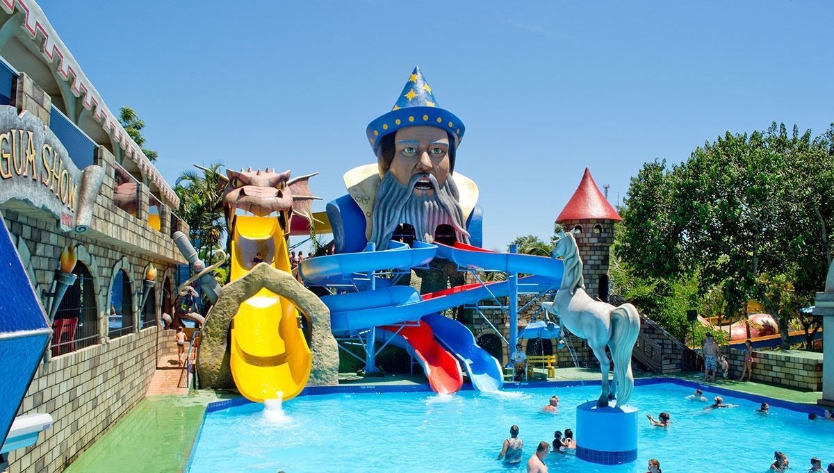 Uma piscina colorida em um parque aquático com uma estrutura gigante temática de mago, toboáguas e pessoas nadando em um dia ensolarado.