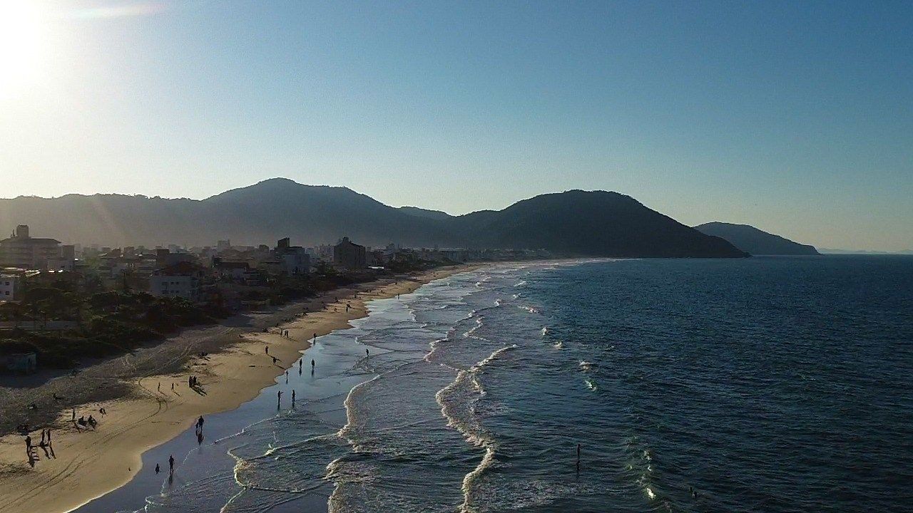 Vista aérea de uma praia ensolarada com ondas quebrando na areia, edifícios ao longo da costa e montanhas ao fundo.