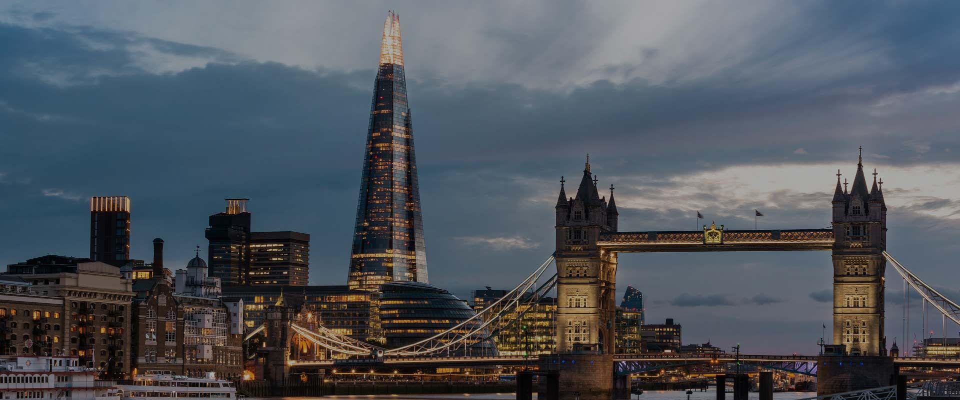O arranha-céu The Shard se ergue acima do horizonte de Londres ao entardecer, com a Tower Bridge iluminada em primeiro plano.