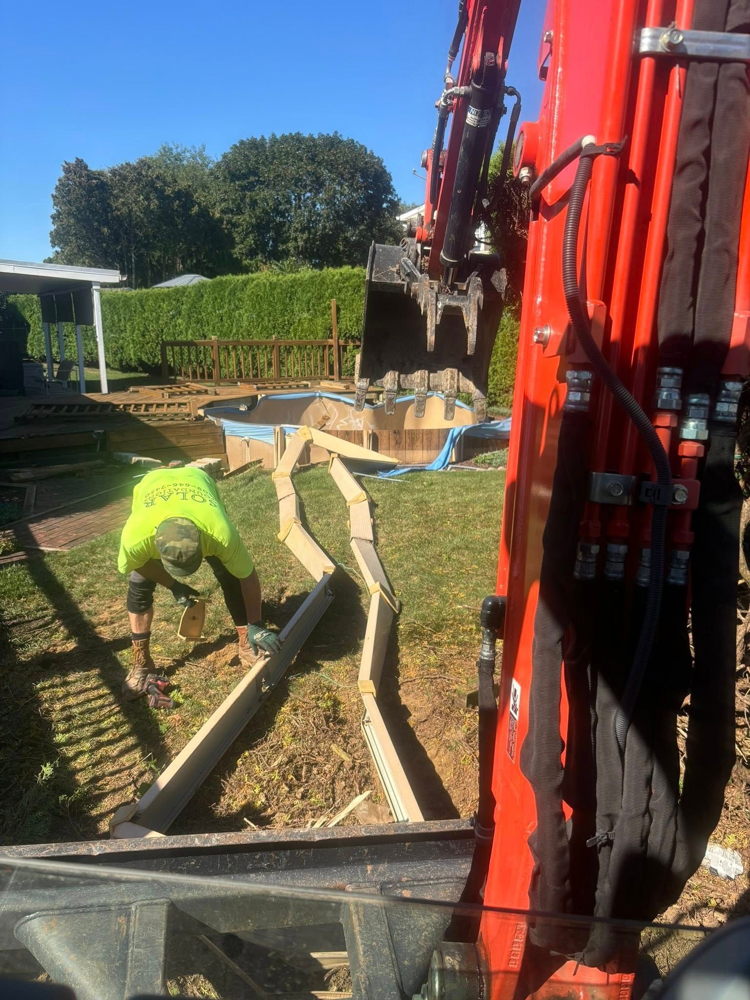 Construction worker using a saw on wooden formwork, excavator in the background. Sunny outdoor setting.