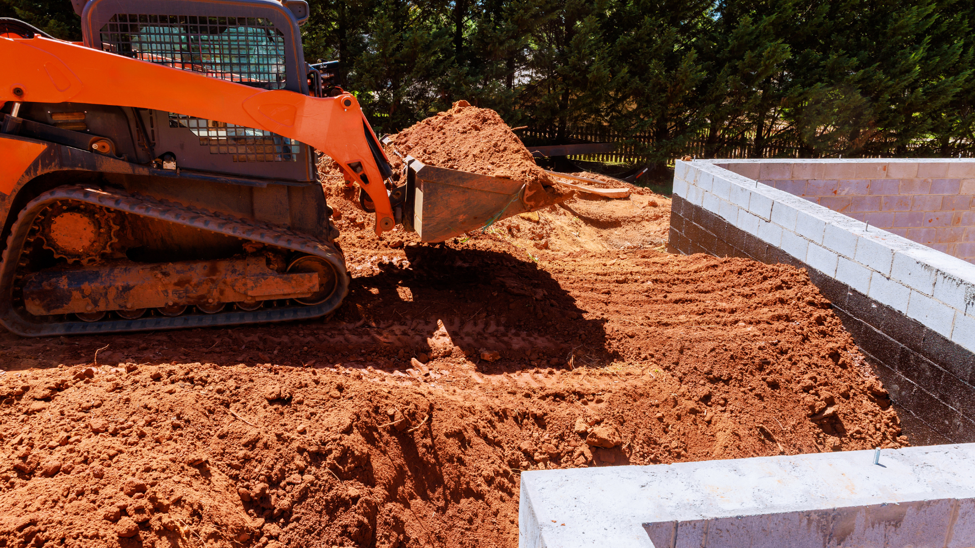 Orange skid steer moving dirt near a concrete foundation.