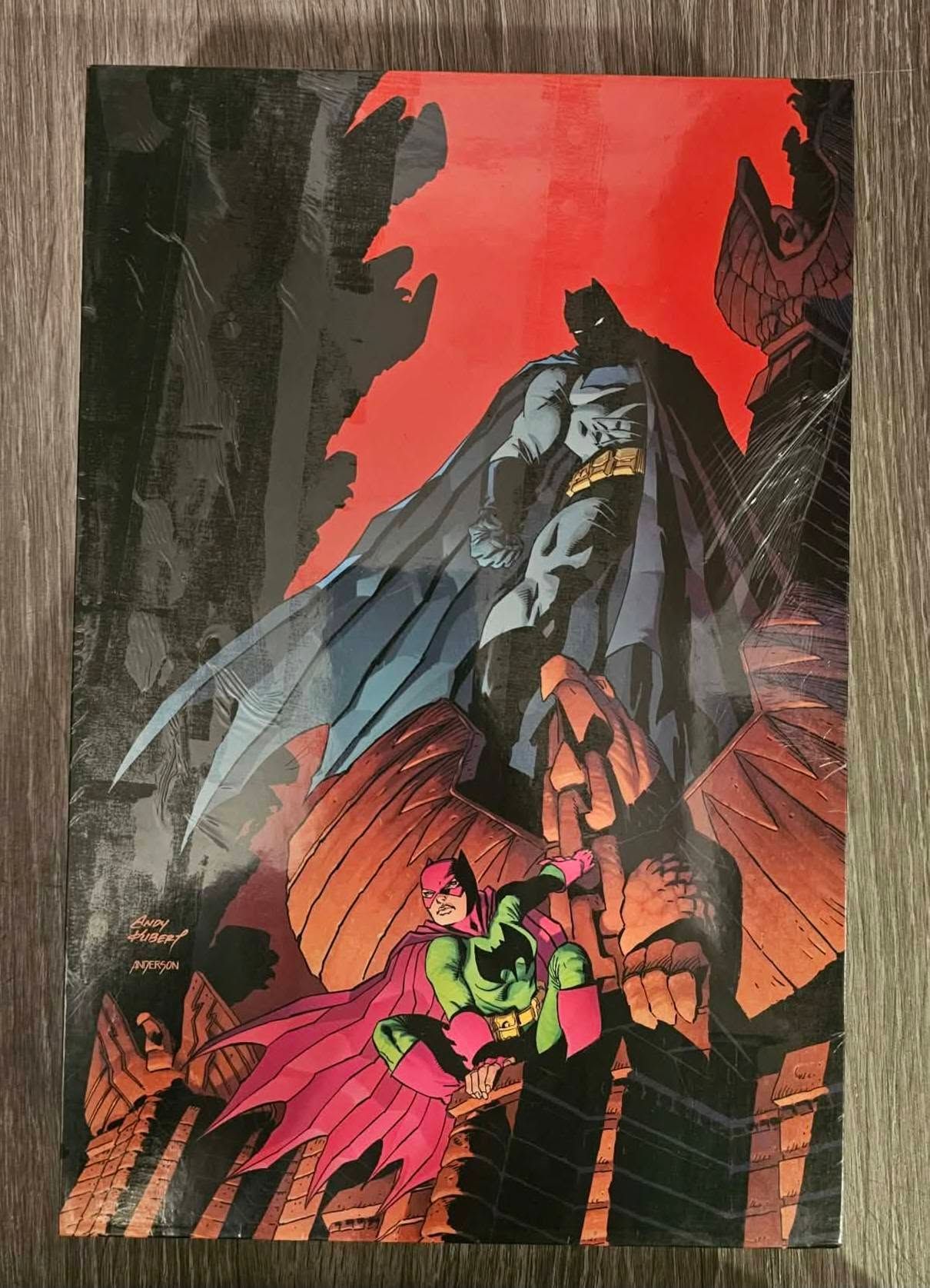 Batman standing on a rocky ledge in a dark cave, with a red sky and glowing orange light below