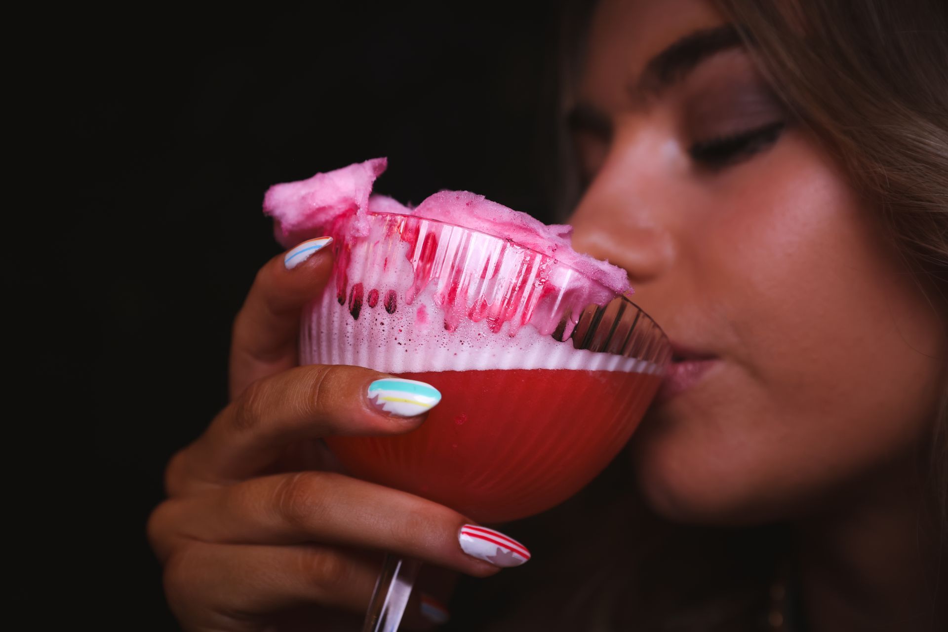 Person sipping a pink cocktail from a martini glass, with red sugar rim and nail art visible