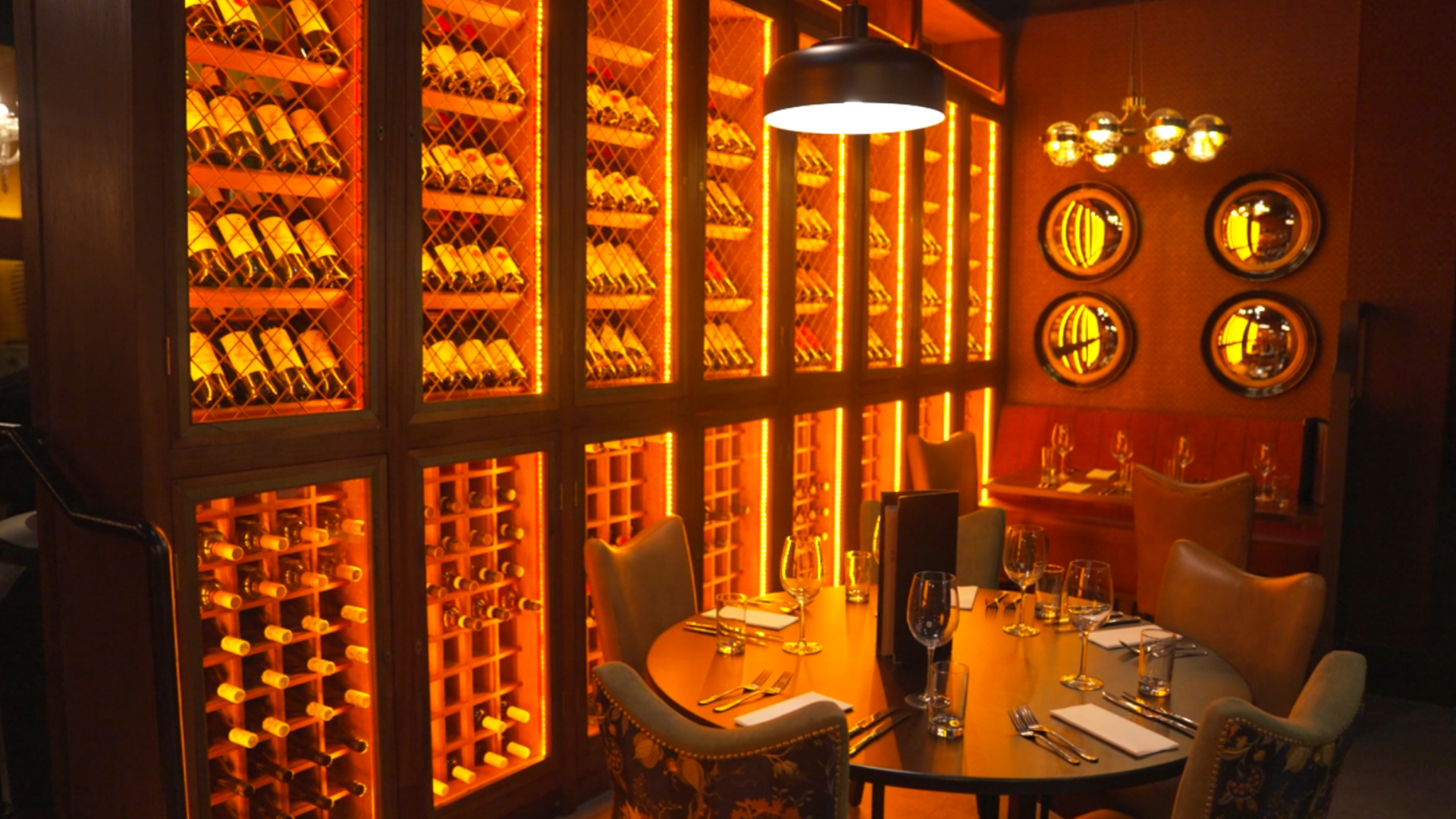 Warmly lit bar interior with amber backlit shelves, round windows, and a small table set for drinks