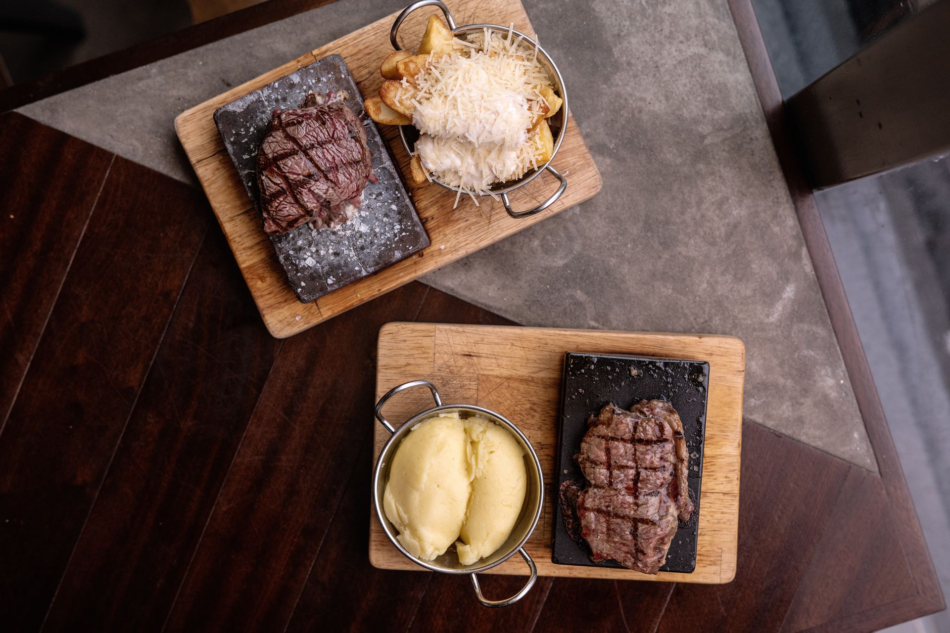 Two wooden plates with grilled steaks and potatoes on a dark table.