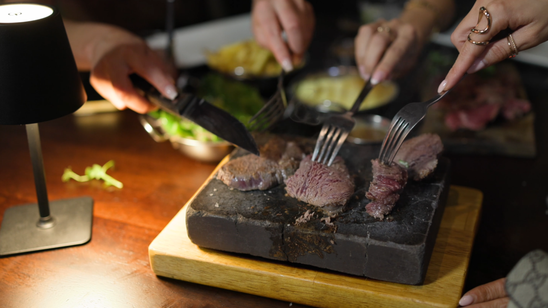 Hands serving sliced steak on a black stone platter at a dimly lit dinner table.