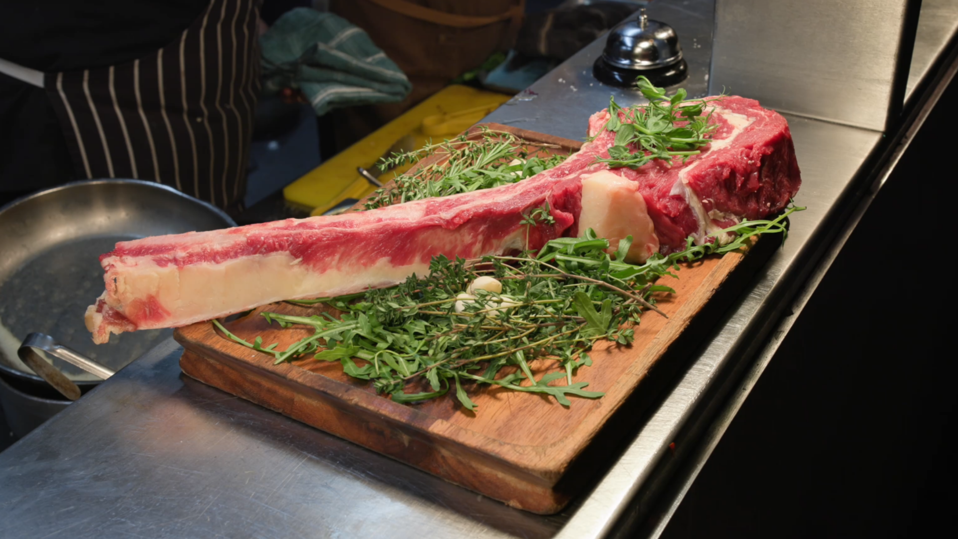 Raw ribeye steak with a long bone on a cutting board, surrounded by fresh herbs in a kitchen