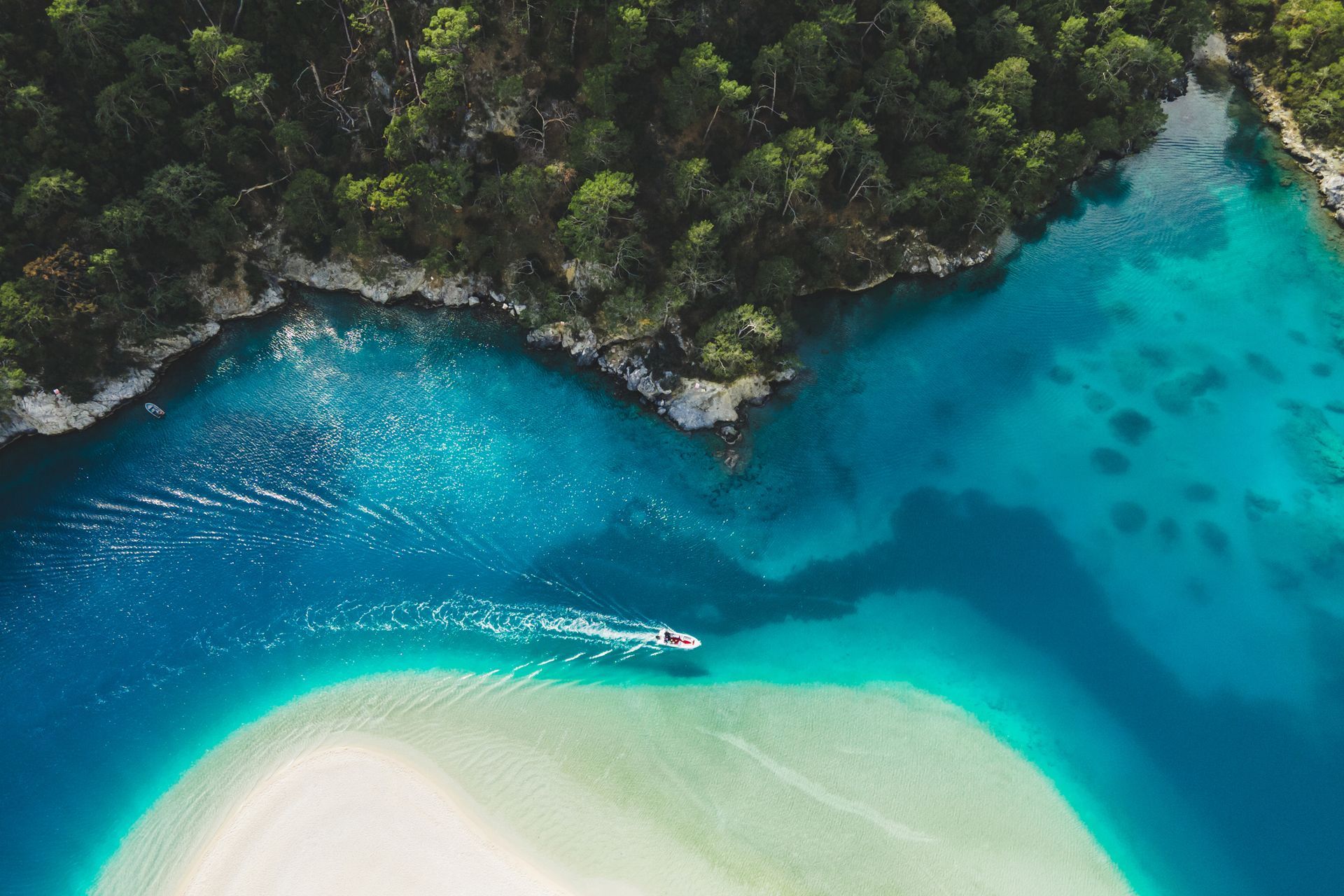 Vista aerea di una laguna turchese con un banco di sabbia e una barca, delimitata da una zona verdeggiante e boscosa.