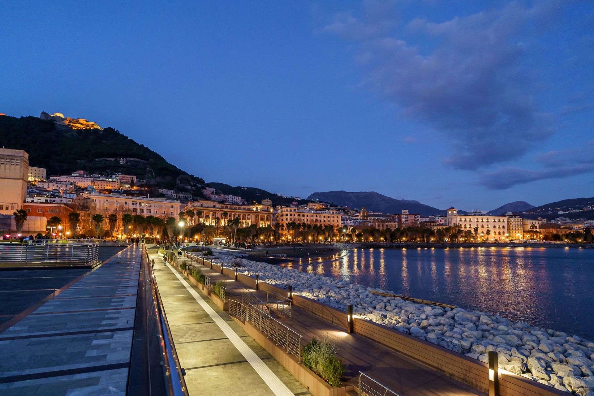 Vista notturna di una città costiera con edifici illuminati che si stagliano contro un cielo blu scuro, una passerella illuminata e uno specchio d'acqua di fronte.