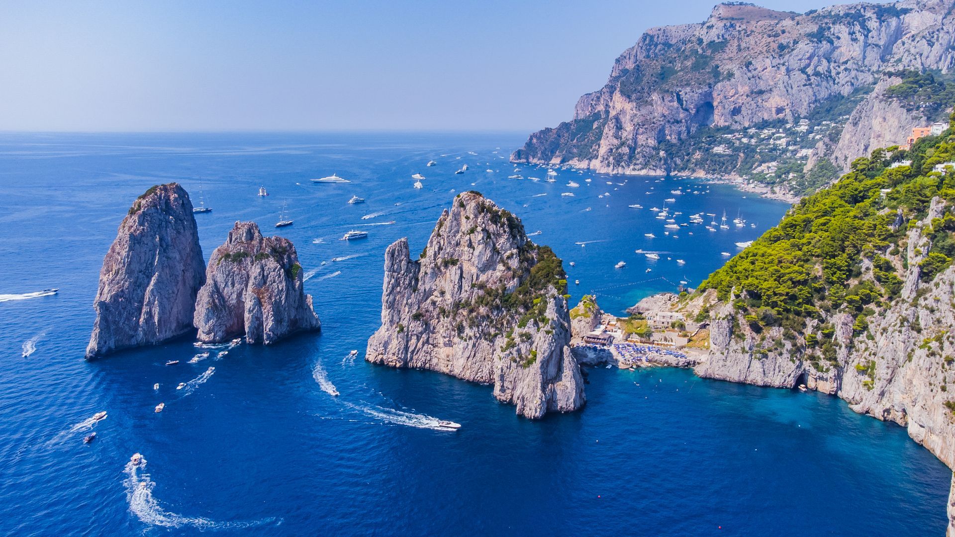 Tre faraglioni si ergono dalle acque azzurre vicino a una costa verde e rocciosa. Nelle vicinanze sono sparse numerose imbarcazioni.