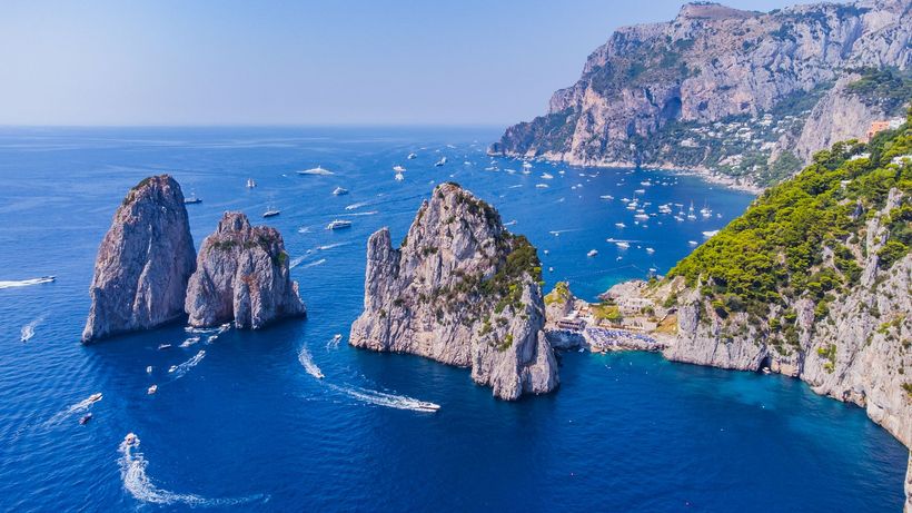 Formazioni rocciose insulari nel blu del Mar Mediterraneo, con barche e scogliere sullo sfondo.