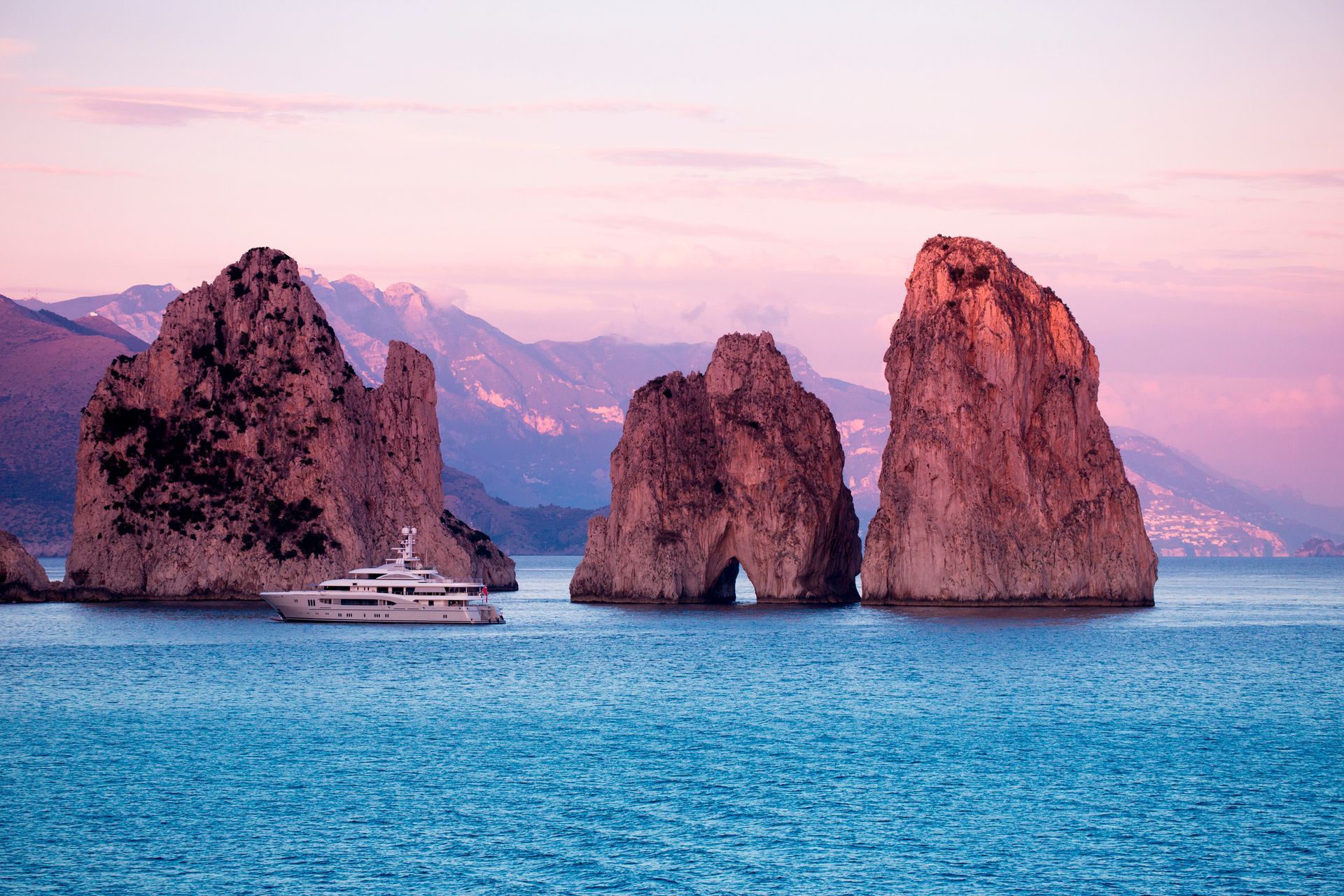 Tre faraglioni si ergono dalle acque turchesi vicino a uno yacht, sotto un cielo al tramonto rosa e viola; Capri, Italia.