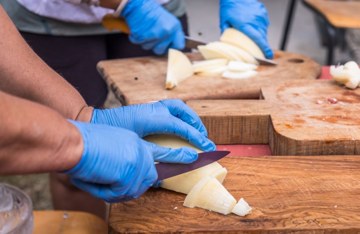 Mani con guanti blu che affettano cibo pallido su taglieri di legno.
