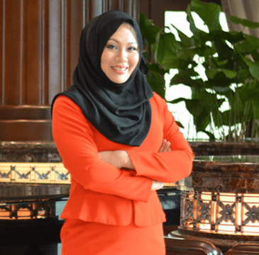 Woman in an orange suit and black hijab smiles, arms crossed, in a formal setting with greenery.