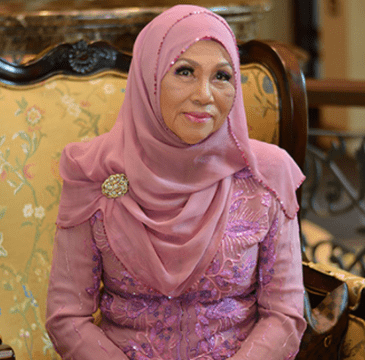 A woman in a pink hijab and dress, seated on a floral-patterned armchair, smiling.