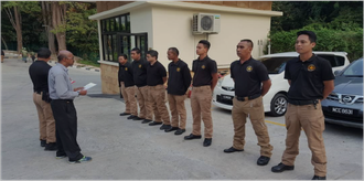 A group of security personnel in uniform standing in line while being addressed by a man holding papers near a building and vehicles.