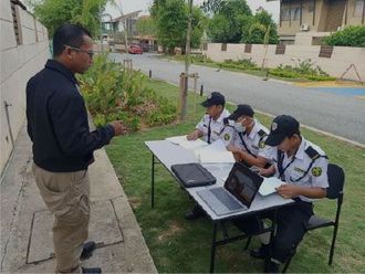 A man talking to three security guards at a table set up outdoors. The guards are checking documents and using a laptop.