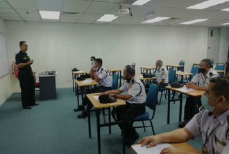 A man in uniform lectures a group of security personnel in a classroom. The students wear masks and sit at desks.
