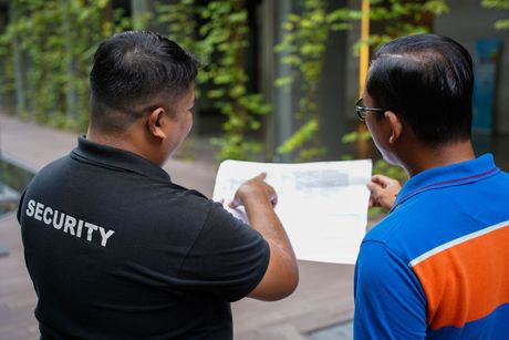 Two men looking at a paper; one is a security guard in a black shirt, pointing, and the other in a blue polo shirt. They are outdoors.