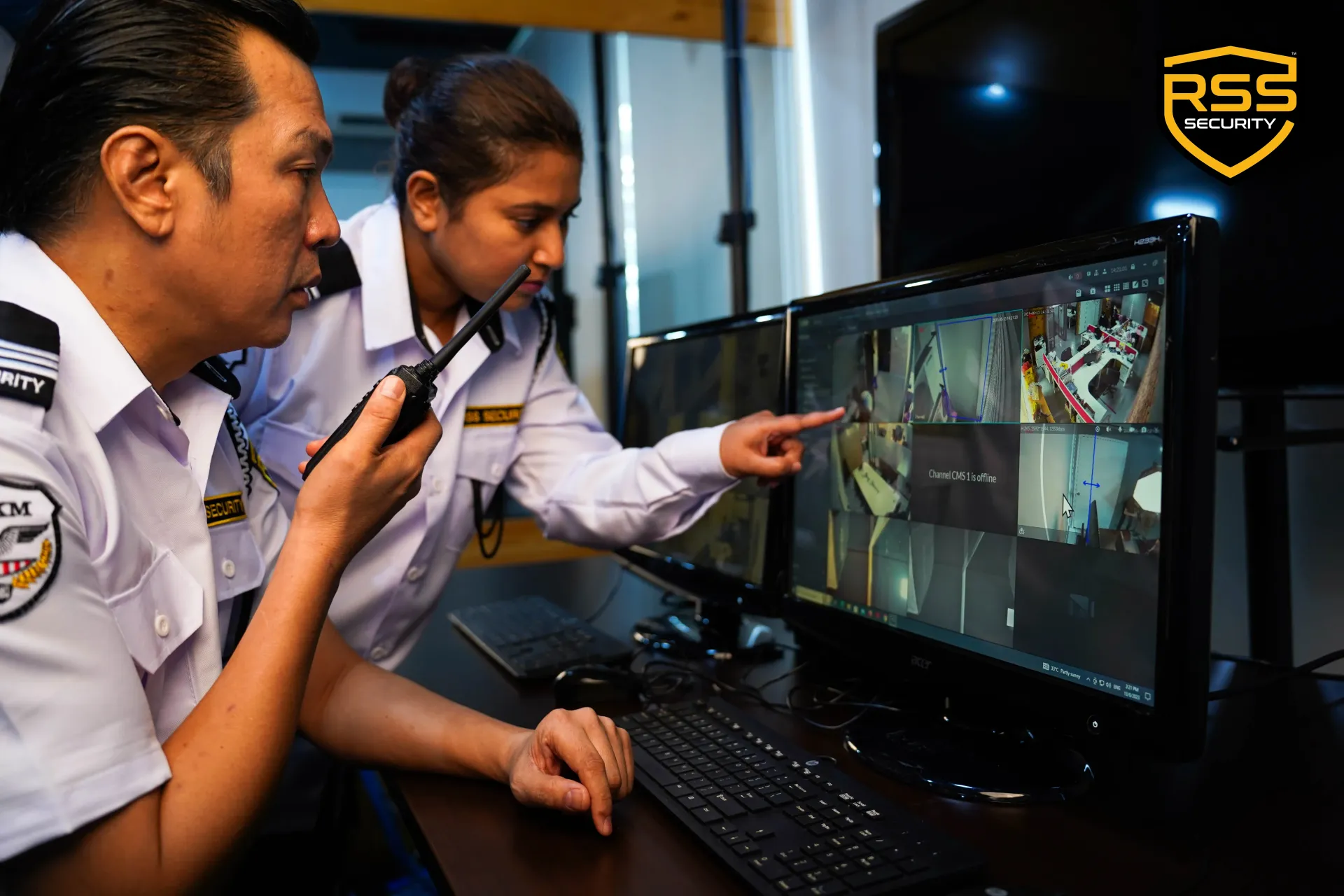 Two security staff in white uniforms monitor surveillance footage on a computer screen.
