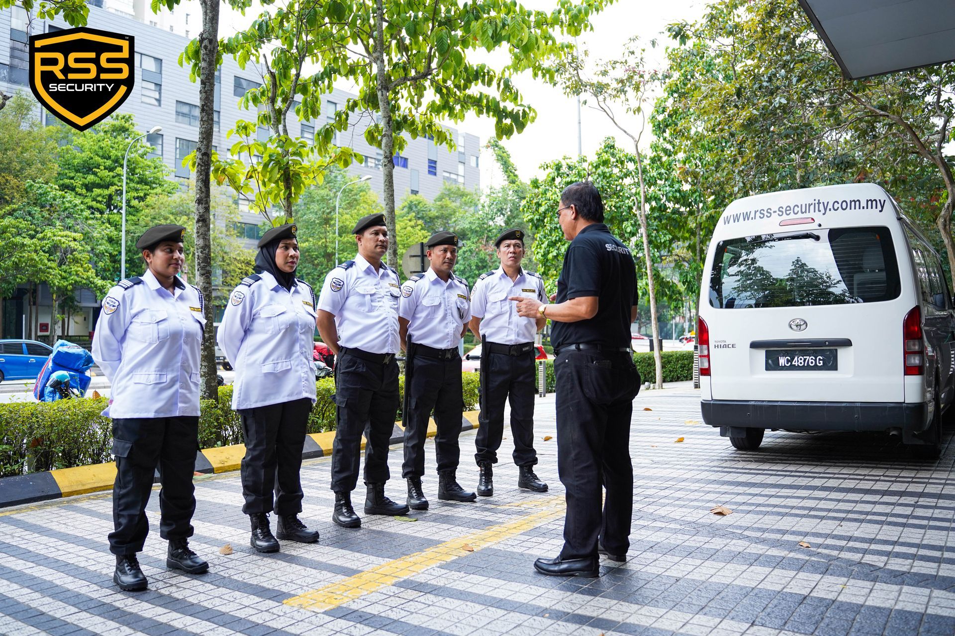 A supervisor in a black uniform briefing four security personnel in white shirts and black trousers in an outdoor lot.