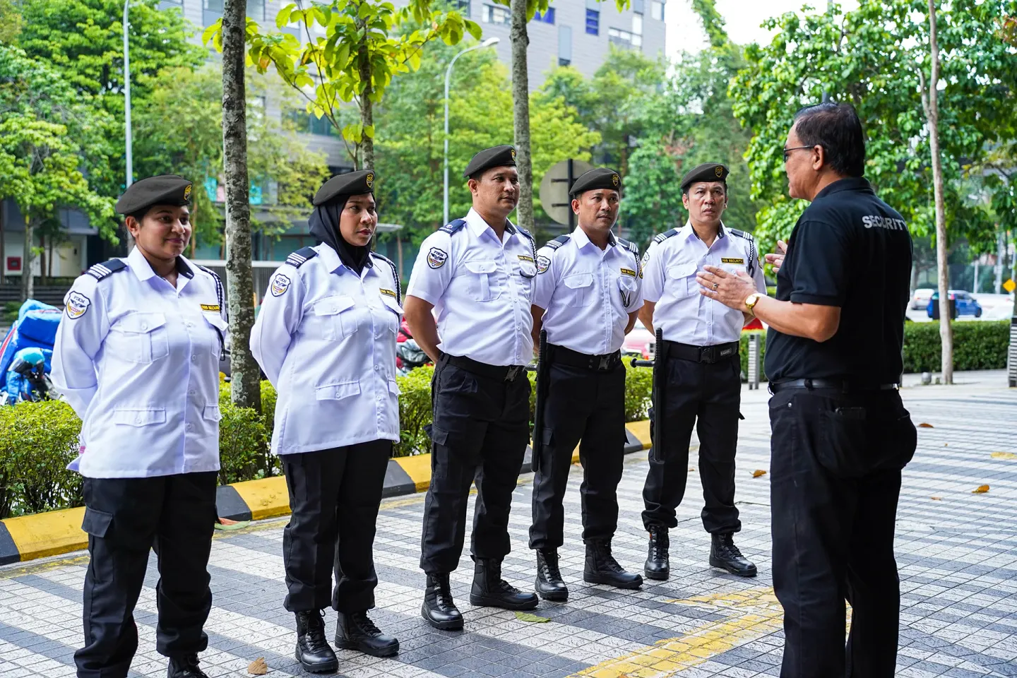 A group of security personnel in blue berets and uniforms listen to a man in black, outdoors near a building.