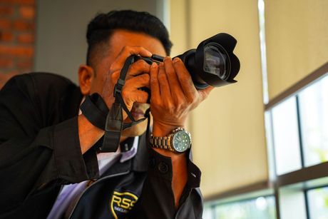 A person with dark skin, holding a black camera to their face, taking a photo. They wear a watch, black shirt, and are indoors near a window.