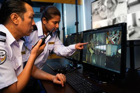 Two security personnel, a man and woman, monitor security cameras at a control station. The woman points at the screen while the man holds a walkie-talkie.