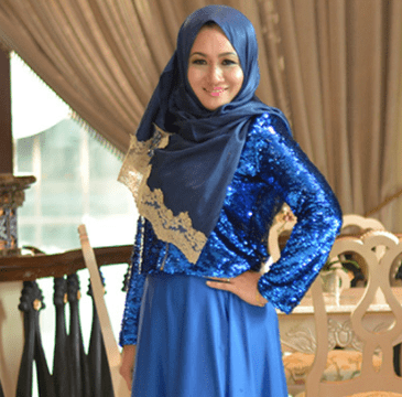 Woman in a blue sequined jacket and hijab smiles, posing indoors.