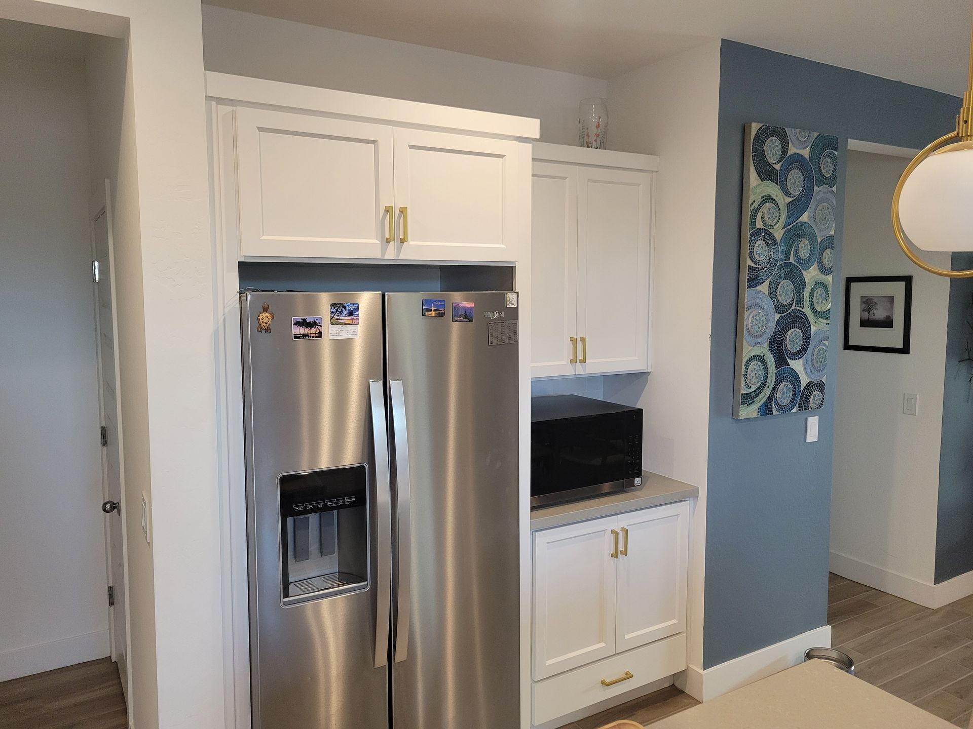White kitchen cabinets surround a stainless steel refrigerator and microwave. A painting hangs on a blue wall.